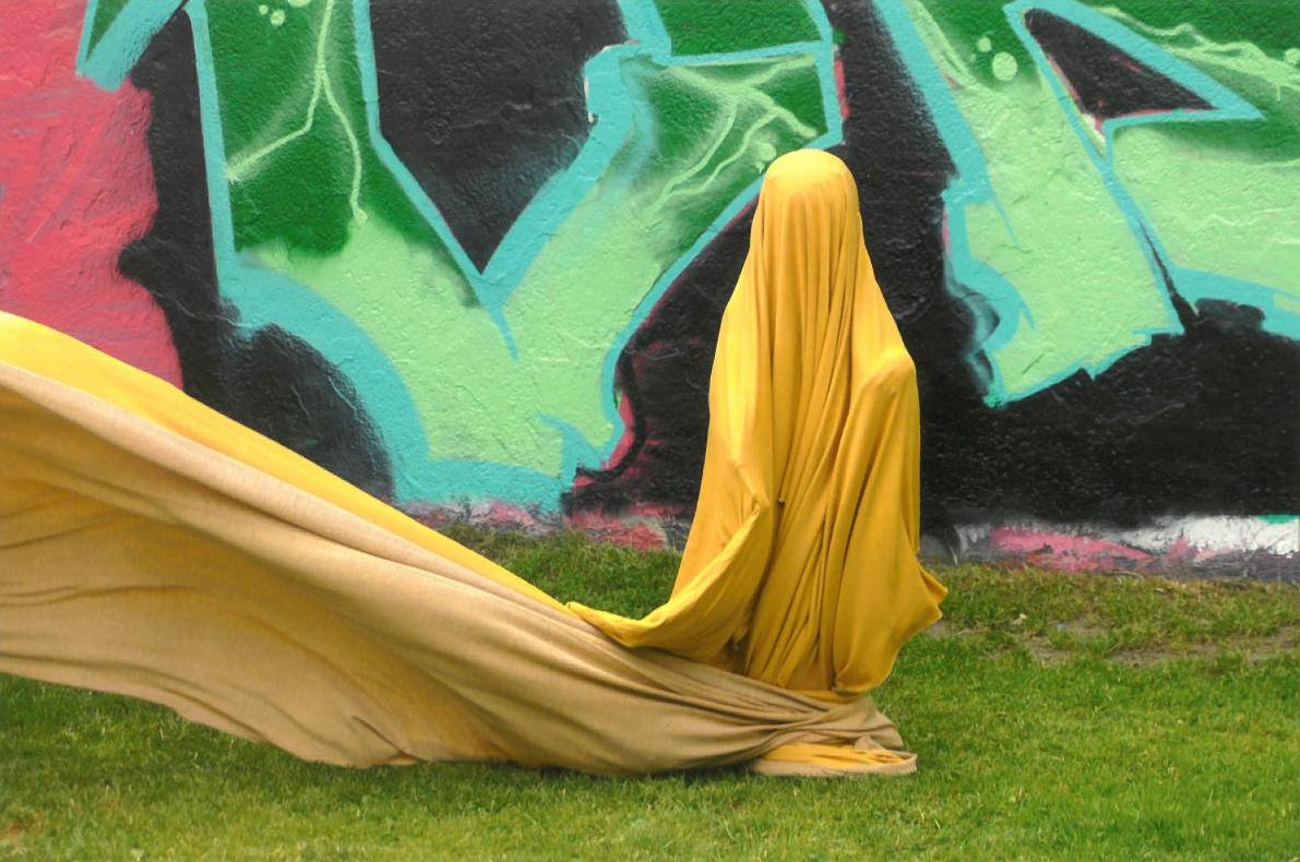 Ein mit schwingendem gelben Tuch verhülltes Kind vor einer bunten Wand. Foto: Katrin Strazzeri