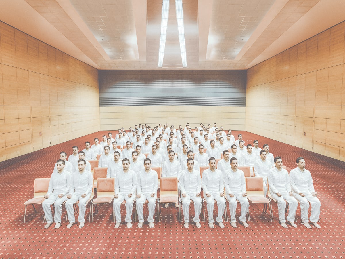 In einem kahlen Raum mit orange-braunem Fußboden und holzfarben getäfelten Wänden sitzen auf schlichten Bänken viele weißgekleidete Männer in geordneten Reihen. Das Bild heißt "Esotericist Seminar". Foto: Klaus Pichler