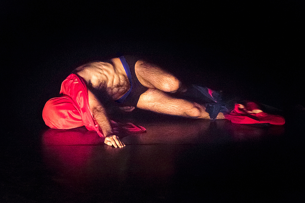 Ein Tänzer mit rotem Tuch über dem Kopf und blauer Hose tanzt liegend auf der Erde 