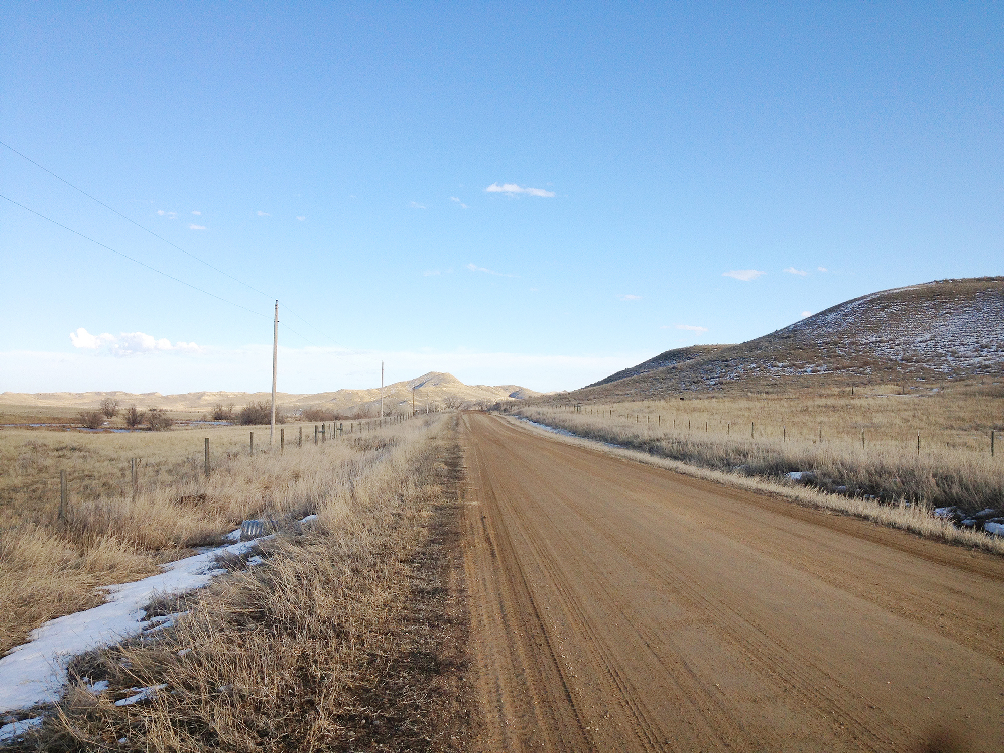 Die Straße Ulm - Ucross. Die Fotografie von Laurie Schwartz zeigt eine lange, braune Straße in Steppenlandschaft, Schneereste am Wegesrand. Keine Menschenseele ist zu sehen: Der Weg in die Geisterstadt Ulm, Wyoming