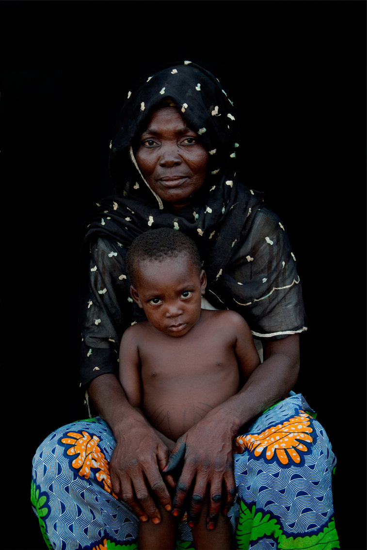 Frau mit dunklem, gepunkteten Kopftuch und buntem Rock mit einem kleinen Kind zwischen ihren Knien vor schwarzem Hintergrund