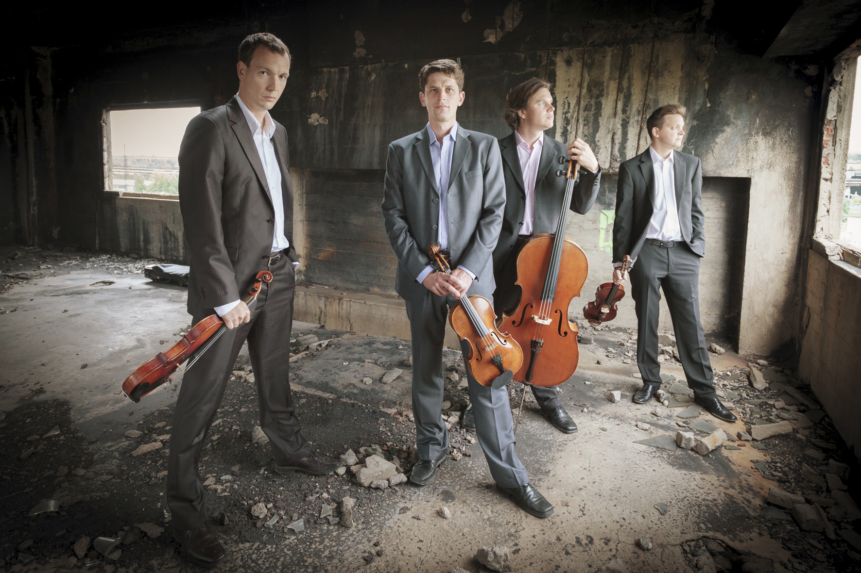 Vier junge Herren in grauen Anzügen, jeder mit seinem Streichinstrument in der Hand, stehen in einem betongrauen Raum. Das Bennewitz-Quartett. Foto: Kamil Ghais