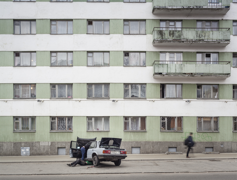 Peter Bialobrzeski: Minsk Diary. In front of a dreary, bare block, a person lies under a white car, trunk, hood and driver's door are open. Another passer-by walks past the house.