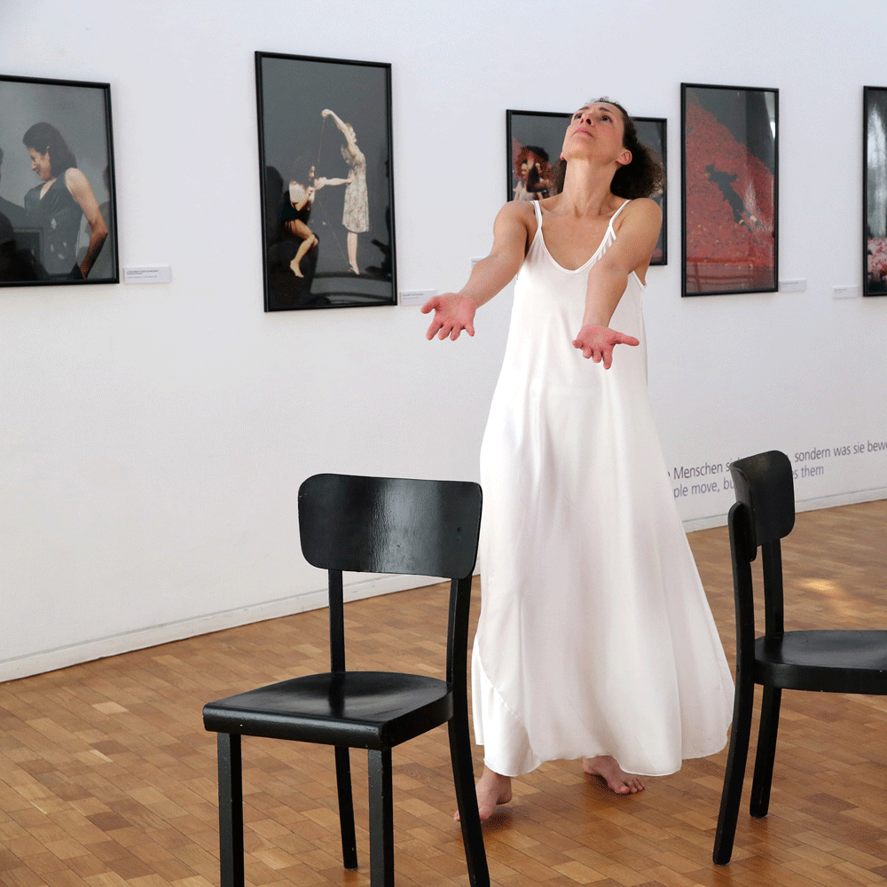 Tänzerin in langem, weißen Kleid tanzt zwischen zwei schwarzen Stühlen bei der Eröffnung der Ausstellung Getanzte Augenblicke. Pina Bausch fotografiert von Ursula Kaufmann im Stadthaus Ulm