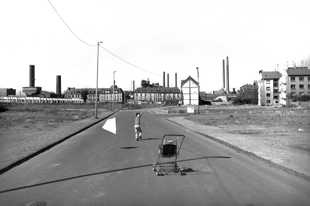 Kind mit Drache auf leerer Straße, im Vordergrund ein Kinderwagen, im Hintergrund Stadtrand