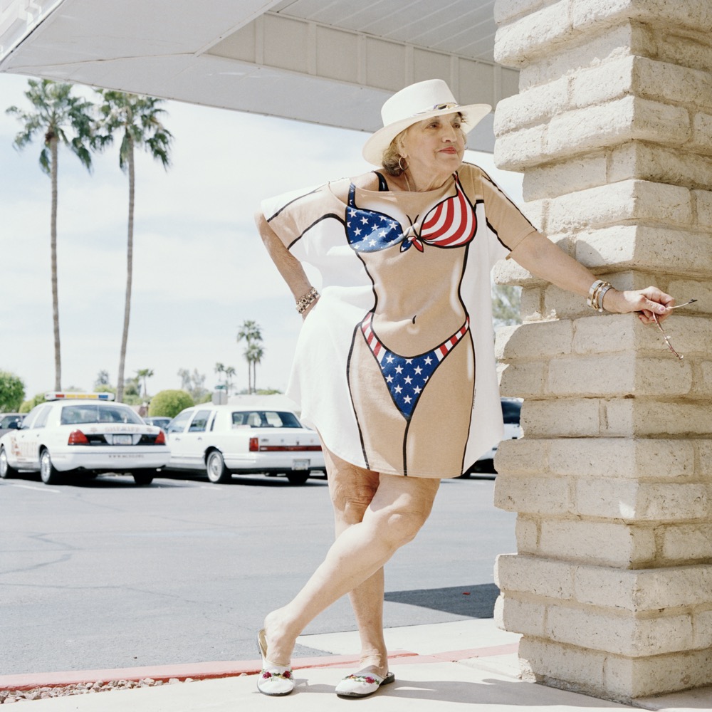 Eine Frau mit Plastikschürze, darauf Bikini in amerikanischen Farben gemalt, steht an Hausecke