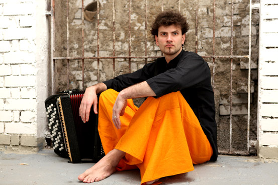 Jüngerer Mann mit schwarzem Oberteil und orangefarbener Hose sitzt mit seinem Akkordeon vor einer Gitterwand auf dem Boden